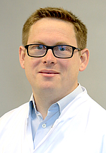 Porträt eines Mannes mit kurzen Haaren und Brille in weißem Hemd vor neutralem Hintergrund