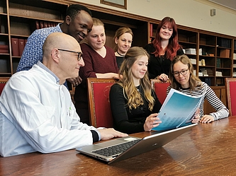 Gruppe von sieben Personen sitzt und steht um einen Tisch mit offenem Laptop und hält ein blaues Dokument in einem Raum mit Bücherregalen im Hintergrund.