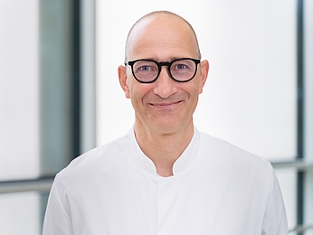 Porträt eines lächelnden Mannes mit Brille und weißem Hemd vor hellem, unscharfem Hintergrund