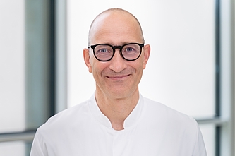 Porträt eines lächelnden Mannes mit Brille und weißem Hemd vor hellem, unscharfem Hintergrund