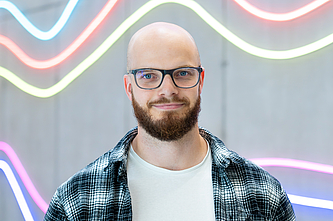 Mann mit Brille, Bart und kariertem Hemd vor einer Wand mit bunten, wellenförmigen Neonlichtern