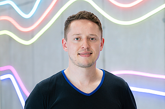 Junger Mann mit kurzem Haar und schwarzem T-Shirt vor einer Wand mit bunten, geschwungenen Neonlichtern